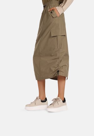 Gonna midi khaki con cintura elastica, tasche laterali e orlo regolabile; abbinata a sneakers beige con dettagli neri e suola spessa.