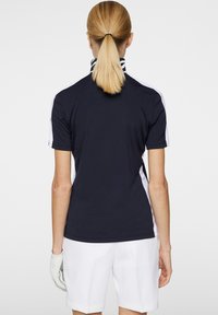 Marinblå polo med vita accenter, korta ärmar och krage. Har en slät textur och en ren design. Vita shorts och en golfhandske.