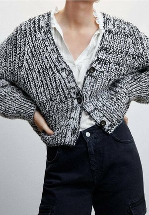 Personne portant un cardigan en tricot noir et blanc sur un chemisier blanc à volants et un pantalon noir taille haute, les mains sur les hanches.