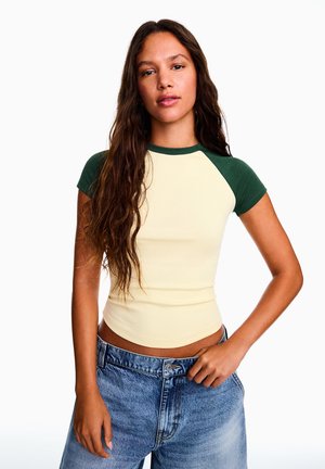 Femme aux longs cheveux ondulés portant un t-shirt raglan jaune et vert et un jean bleu, debout devant un fond blanc uni.