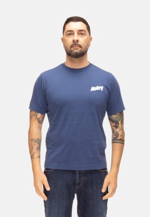 Hombre con cabello corto y oscuro y barba, vistiendo una camiseta azul Aubry y vaqueros, mostrando tatuajes en ambos antebrazos, de pie contra un fondo blanco.