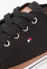 Baskets noires avec lacets noirs, œillets argentés, petit logo rectangulaire rouge, blanc et bleu, et bordure marron au-dessus de la semelle blanche.