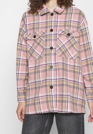 Long-sleeve pink plaid shirt jacket with black buttons and two large front flap pockets, worn over dark jeans.