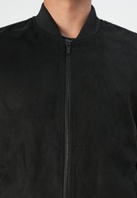 Svart bomberjakke laget av glatt stoff. Har glidelås og ribbestrikket krage. Teksturen ser myk ut med en matt overflate.
