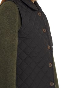 Gilet trapuntato nero con bottoni marroni e colletto. Il gilet è indossato sopra un indumento lavorato a maglia verde scuro.
