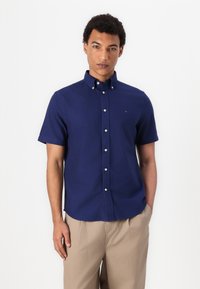 Camicia button-up a maniche corte blu navy in cotone, con colletto classico e un piccolo logo sul petto, abbinata a pantaloni beige.