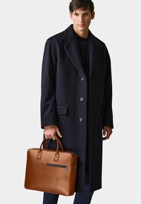 Navy wool overcoat with notched collar, button front. Brown leather briefcase with top handles, zip pocket, and embossed logo detail.