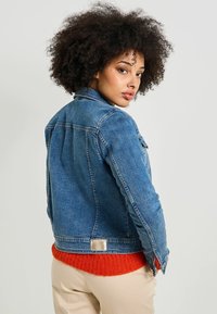 Chaqueta de denim de corte clásico, color azul, con etiqueta de metal plateado en la parte posterior; superpuesta sobre un suéter de punto naranja brillante.