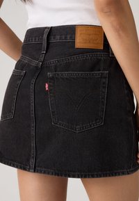 Γυναίκα που φοράει μαύρη φούστα από τζιν Levi’s με πίσω τσέπες, δερμάτινή ετικέτα του brand και λευκό ριμπ αμάνικο τοπ.