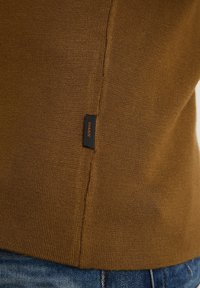 Ourlet de pull marron en maille avec petite étiquette en tissu noir portant la mention "CHASIN", porté par-dessus un jean en denim bleu.