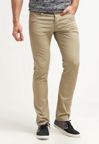 Homme portant un pantalon slim beige, des baskets grises et noires, et une chemise grise à motifs avec une main dans la poche.
