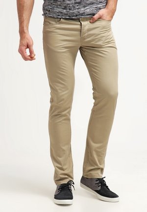 Muž v béžových slim-fit kalhotách, šedých a černých teniskách, a šedé košili s vzorem, s jednou rukou v kapse.