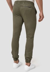 Mann trägt olivgrüne Jogginghosen mit elastischen Bündchen, Reißverschlusstasche hinten und weißen Sneakers, steht vor schlichtem Hintergrund.