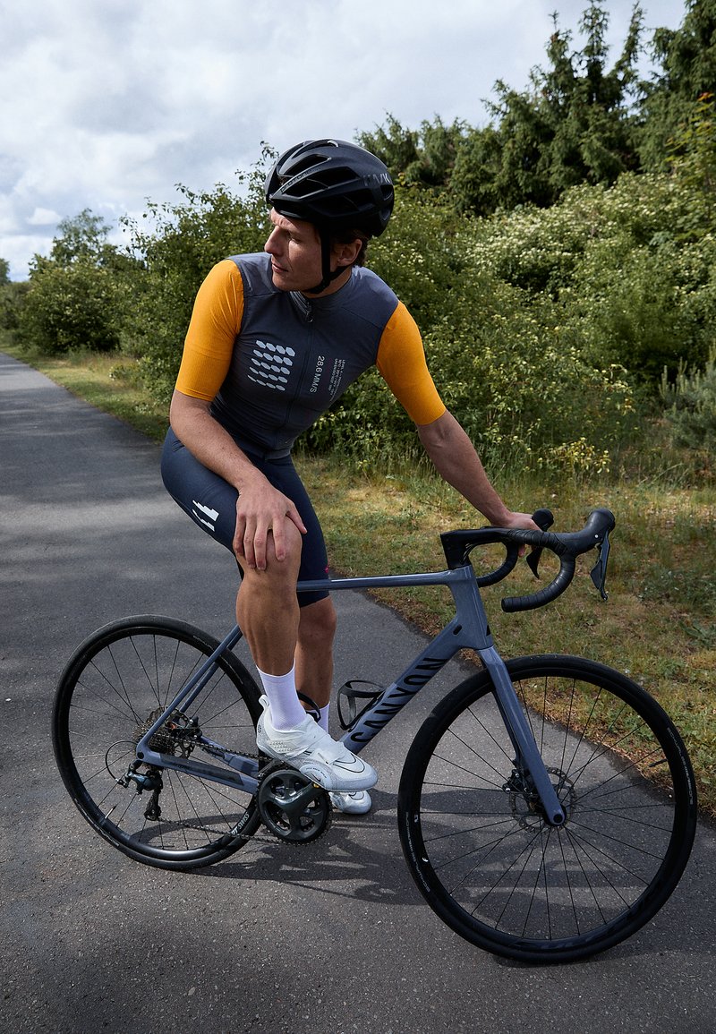 Graues Rennrad mit schlankem Rahmen, schwarzen Rädern und einem Fahrer in einem eng anliegenden grauen und orangefarbenen Trikot. Sneakers und ein schwarzer Helm vervollständigen das Aussehen.