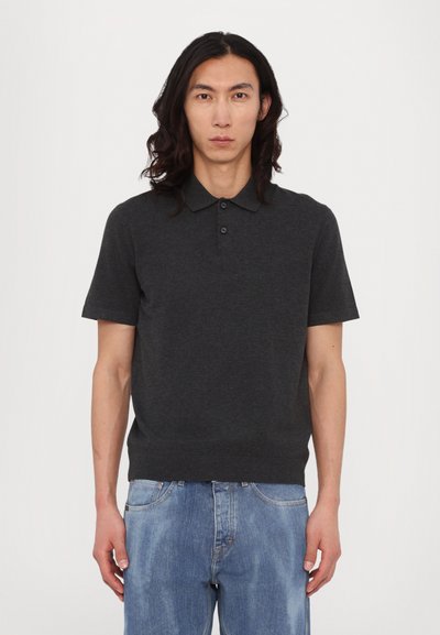 Camisa polo de manga corta en gris oscuro con un collar clásico, cierre de tres botones y un diseño ajustado. Lleva puesta con jeans de mezclilla azul claro.