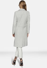 Svetle šedý elegantní kabát s klopovým límcem a rozparkem na zadní straně, spolu se světlými šedými kalhotami. Černé sandály na vysokém podpatku outfit doplní.