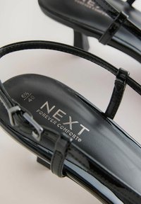 Crne lakirane sandale s niskom petom, s tankim trakama i podstavljenim uloškom. Vidljiv "NEXT FOREVER COMFORT" brend.