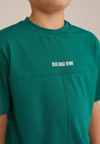 Žalias medvilnės marškinėliai su apvaliu kaklu, trumpomis rankovėmis ir balta "BLUE RIDGE DENIM" užrašu, papuošti kontrastingomis siūlėmis.