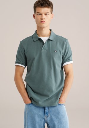 Polo in gedempt groen katoen, met een klassieke kraag, korte mouwen met witte bies, en een klein geborduurd logo op de linkerborst.
