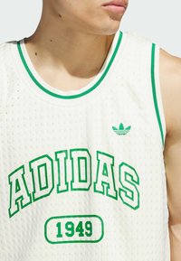 White, sleeveless athletic jersey with green accents, featuring the text "ADIDAS" and "1949." Textured mesh material, small green logo.