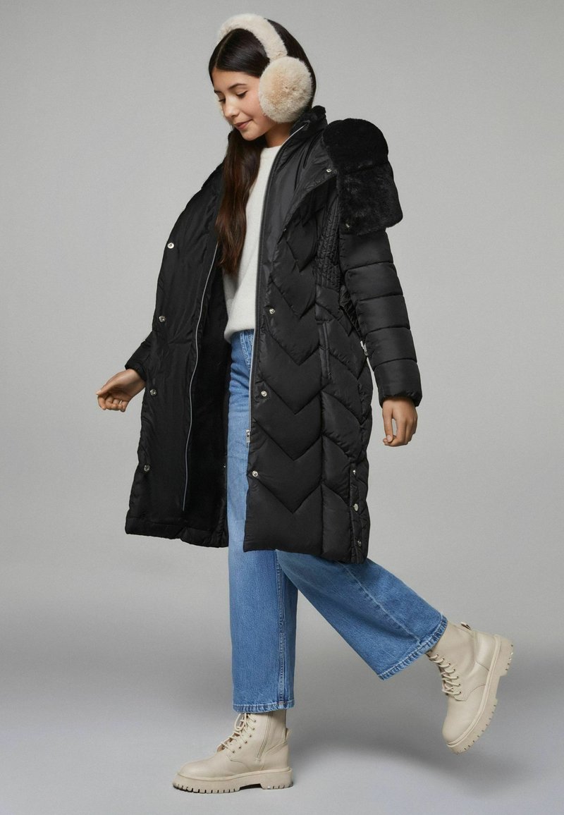 Cappotto lungo imbottito nero con motivo a zigzag, bottoni a pressione e dettagli in pelliccia sintetica. Indossato con jeans blu chiaro e stivali beige con lacci.