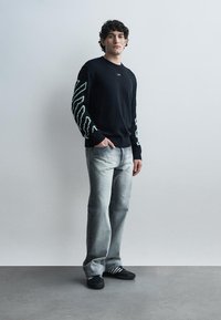 Un jeune homme se tient contre un mur uni, portant un pull noir avec des motifs blancs sur les manches, un jean gris délavé et des baskets noires.