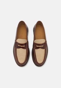 Mocassins en cuir marron avec un panneau en tissu beige, présentant des coutures décoratives et un petit accent en cuir sur le devant.