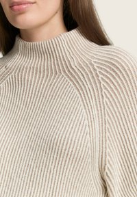 Sweater tricoté en crème et beige avec une texture en côtes verticales, col haut et tissu doux. Les détails de couture ajoutent de la dimension au design.