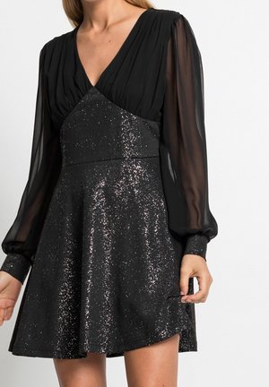 Donna che indossa un vestito nero scintillante con maniche lunghe trasparenti e scollatura profonda a V, mentre tiene l'orlo della gonna.