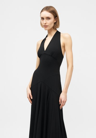 Femme aux cheveux courts et blonds portant une longue robe noire sans manches à col halter, sur un fond clair uni.