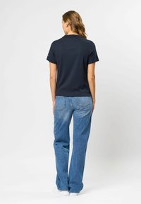 Kvinne stående med ryggen mot, iført en svart kortermet topp, blå denimjeans og hvite joggesko mot en ensfarget bakgrunn.