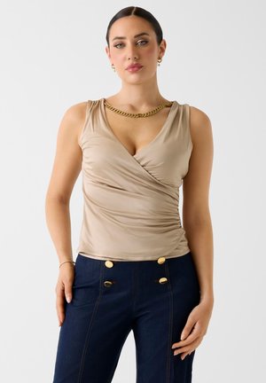 Frau mit dunklem Haar, die ein ärmelloses, beiges Wickeloberteil und marineblaue, hoch taillierte Hosen mit goldenen Knöpfen trägt, dazu eine goldene Kettenhalskette und Ohrringe.