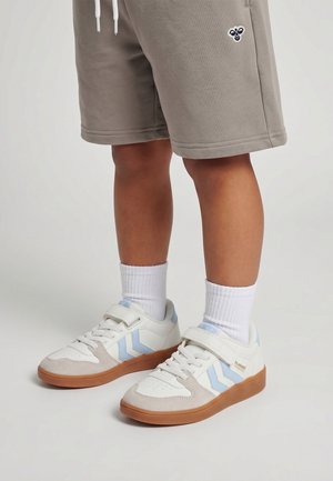 Kind draagt beige korte broek met bijenlogo, witte sokken en witte sneakers met lichtblauwe chevronstrepen en rubberen zolen.