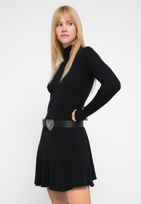 Čierne rebrované pletené šaty s vysokým golierom a rozšírenou sukňou, ozdobené čiernym opaskom so striebornou prackou v tvare srdca.