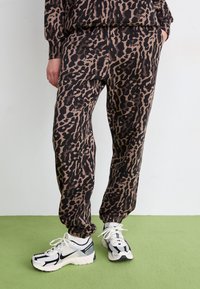 Joggerji z leopardjim vzorcem v črni in rjavi barvi z elastičnim pasom in manšetami na gležnjih, v kombinaciji z belimi in črnimi športnimi čevlji.