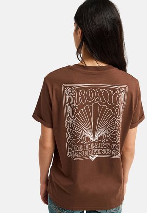 Person mit langen dunklen Haaren trägt ein braunes Roxy-T-Shirt mit Muschel- und Blumenmotiv und dem Text "The heart of surfing" auf dem Rücken.
