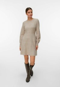 Strikket beige kjole med rund halsudskæring og lange ærmer, der vidner ved taljen. Sammen med knæhøje brune støvler. Glat tekstur.
