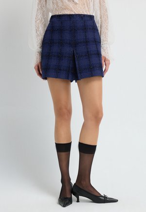 Marineblau karierten Shorts aus strukturiertem Stoff, mit einem Schlitz vorne und schwarzen Akzenten. Dazu wird ein Paar halbtransparente schwarze knielange Strümpfe und spitze Schuhe getragen.