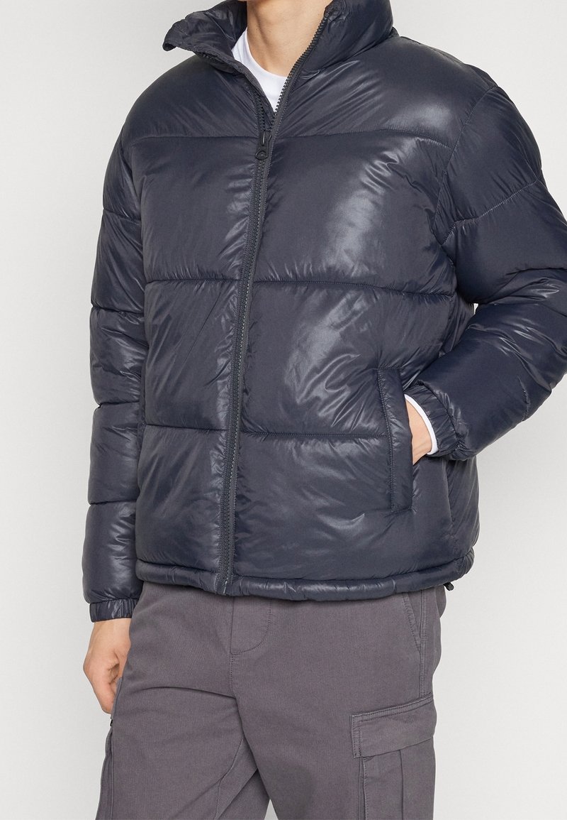 Donkergrijze donzen jas met glanzende afwerking, een hoge kraag, ritssluiting en zijzakken. Voorzien van een horizontaal doorgestikt patroon en elastische manchetten.