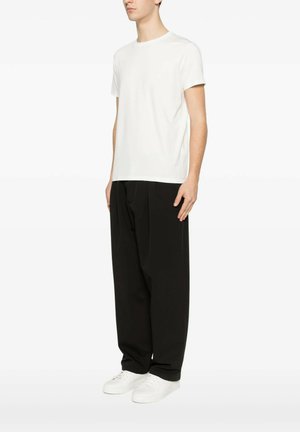 Uomo che indossa una t-shirt bianca semplice a maniche corte, pantaloni neri larghi e scarpe da ginnastica bianche, in piedi davanti a uno sfondo bianco uniforme.