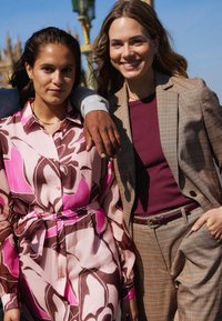 Dva jedinci stojí společně, jeden v růžovo-hnědé květinové blůze s knoflíky, druhý v hnědém kostkovaném obleku s opaskem.