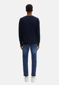 Marineblauwe trui met ronde hals, geribbelde manchetten en zoom, gecombineerd met blauwe spijkerbroeken met een subtiel ontwerp op de achterzak en witte sneakers.