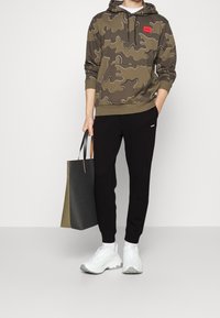 Olijfgroene camouflage hoodie met een rode logopatch, gecombineerd met zwarte broek. Witte sneakers met grijze zolen en een zwarte tote bag maken de outfit compleet.