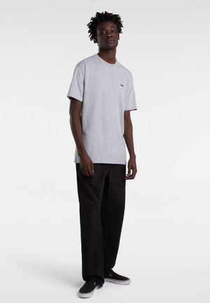 Mand stående iført en lysgrå Vans t-shirt, sorte løse bukser og sorte slip-on sneakers med hvide såler mod en hvid baggrund.