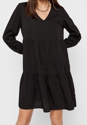 Kobieta ubrana w luźną czarną sukienkę z długim rękawem, dekoltem w serek i warstwową spódnicą, udekorowaną grubym złotym naszyjnikiem łańcuchowym.