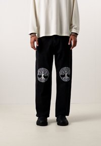 Zwarte broek met geborduurde boomontwerpen op de knieën, gecombineerd met een crèmekleurig shirt met lange mouwen en zwarte schoenen.