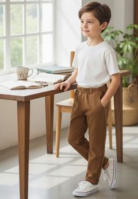 Biała koszulka z krótkim rękawem, brązowe spodnie z elastycznym pasem, białe sneakersy z granatowymi akcentami. Stoi przy stole z książkami i kubkiem.