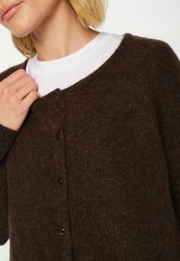 Brūns strukturēts kardigāns, izgatavots no mīksta materiāla, ar apaļu izgriezumu, garām piedurknēm un brūnām pogām priekšpusē.