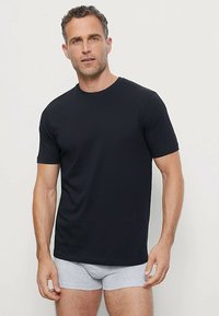 Zwart T-shirt met korte mouwen, gemaakt van glad katoen, met een ronde hals. Draagt over grijze slip, model staat tegen een effen achtergrond.