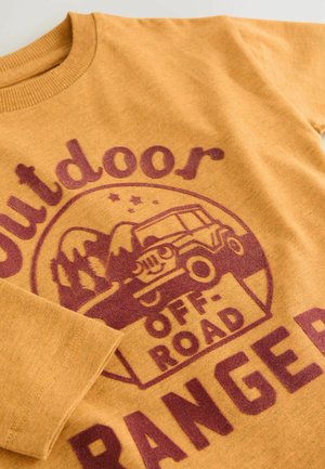 Sweat-shirt de couleur moutarde avec un col côtelé. Présente un imprime maron de montagnes et d'un véhicule tout-terrain vintage, avec le texte "Outdoor Ranger."